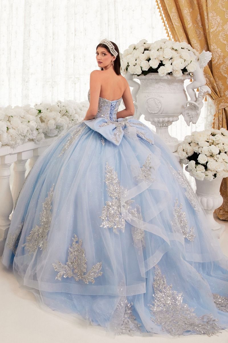 A woman in the Ladivine Strapless Quinceanera Dress 15715 with silver embellishments poses before grand white flower arrangements and gold curtains, looking over her shoulder.